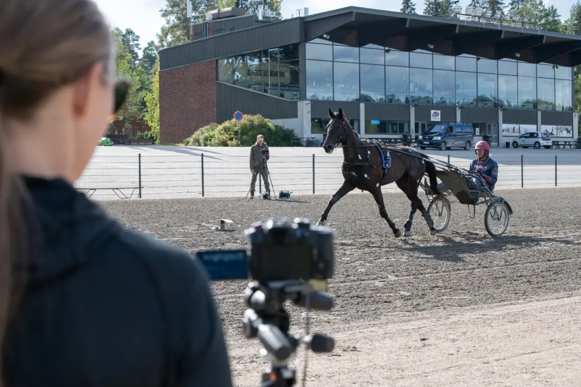 Ravihevonen, ohjastaja ja kärryt juoksemassa Killerin raviradalla. Radan molemmin puolin tutkija kuvaa videokameralla. Taustalla näkyy Killerin katsomorakennus.